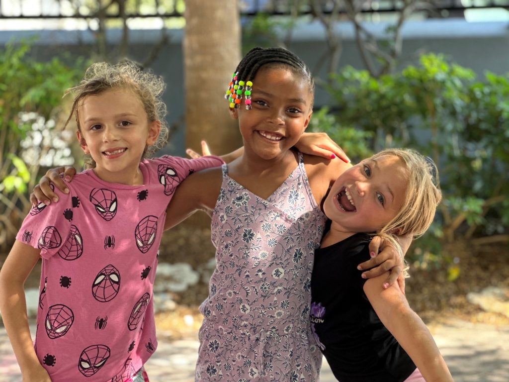 Three young girls from Girls Inc. of Sarasota County smile at camera outside with tree behind them.