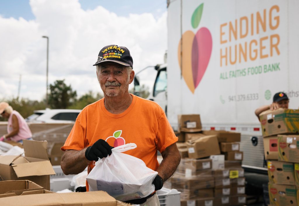 Man standing outside with bag of food in hands wearing orange All Faiths Food Bank shirt.