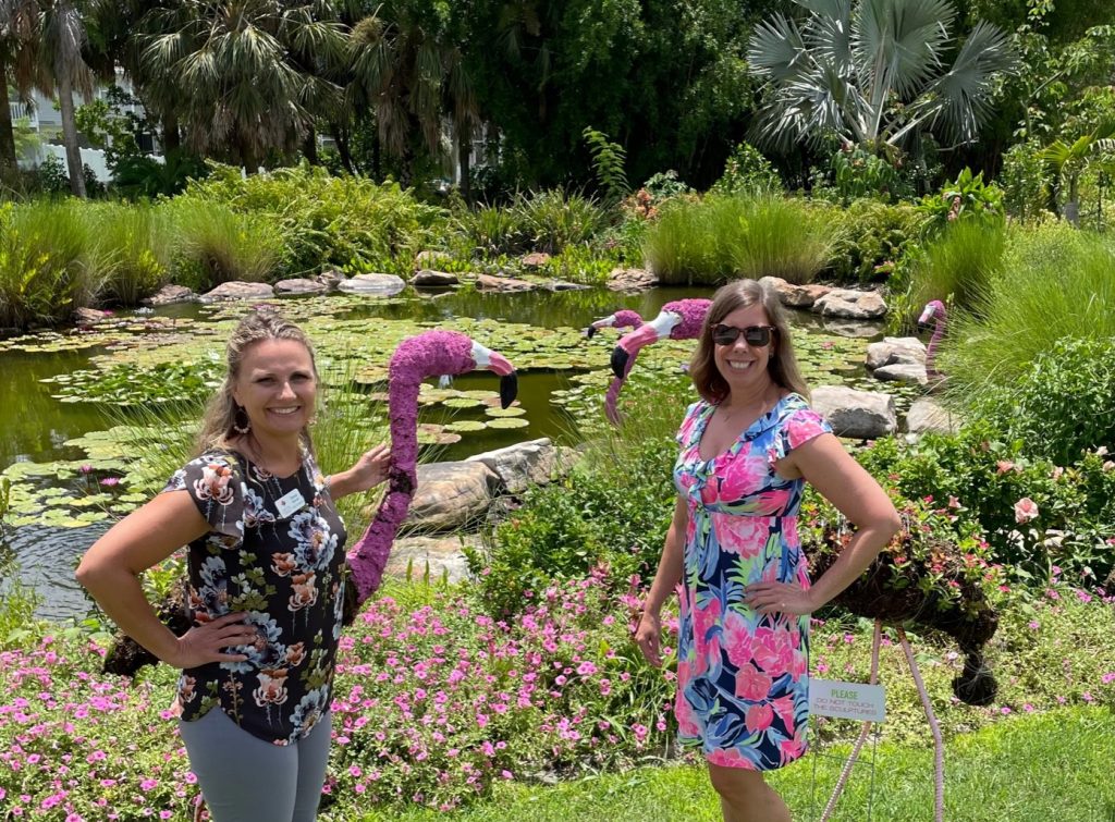 Two women standing outside in botanical gardens with two crafted flamingos.