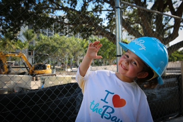 One small child wearing I love The Bay shirt and helmet waves at camera.