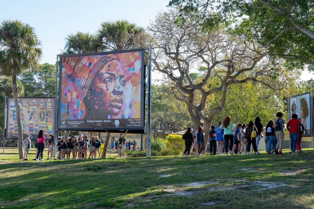 Large paintings outdoors in art exhibit with students gathering around to view them.