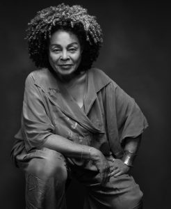 Black woman sits with her elbow on her knee smiling at camera.