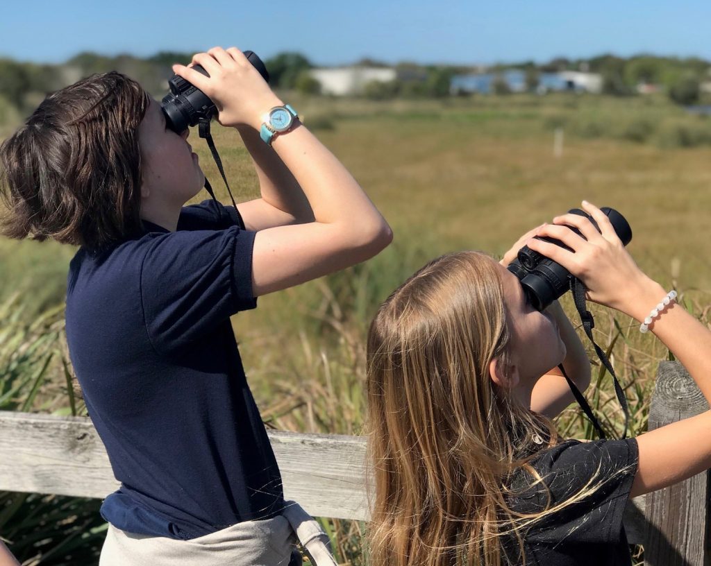 Two children in blue shirts use binoculars outside to look at the wildlife around them.